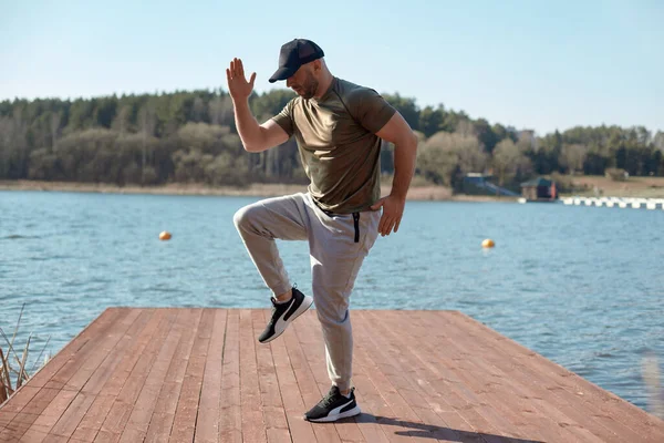 A strong athletic man in sports clothes does exercises on the pier on the lake. A man does sports. Sports, fitness, lifestyle