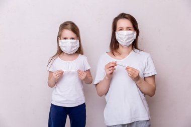 Koronavirüs karantinası kavramı. Genç bir kadın ve tıbbi maskeli kızı evde tecritte. Anne kızının sıcaklığını ölçer. Sağlık ve Tıp Teması.