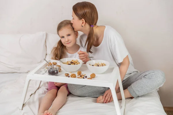 Happy mom and daughter have healthy breakfast on bed in a light bedroom on a sunny morning. Mom kisses daughter. Healthy food and family relationships concept. Good mood.