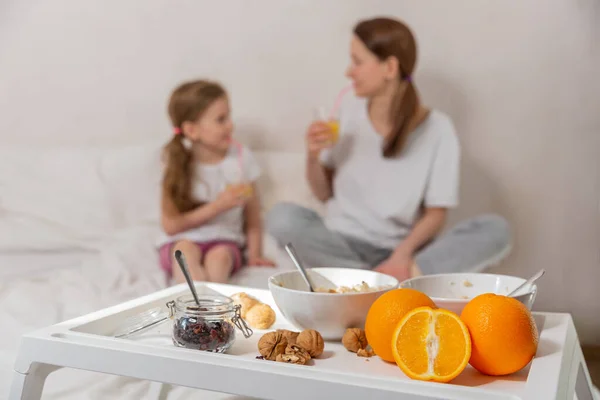 Happy mom and daughter have healthy breakfast on bed in a light bedroom on a sunny morning. A table with breakfast in the foreground and mom and daughter in defocus. Healthy food concept. Good mood.