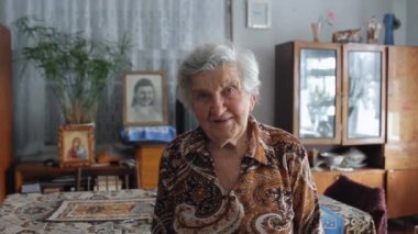Kıdemli kadın kameraya bakıyor, eski Sovyetler Birliği dairesinde poz veriyor, portreyi kapatıyor.