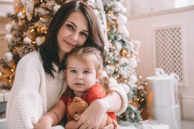 Mutlu Noeller ve mutlu tatiller. Neşeli anne ve tatlı kızı içeride Noel ağacının yanında eğleniyorlar, kameraya gülümsüyorlar..