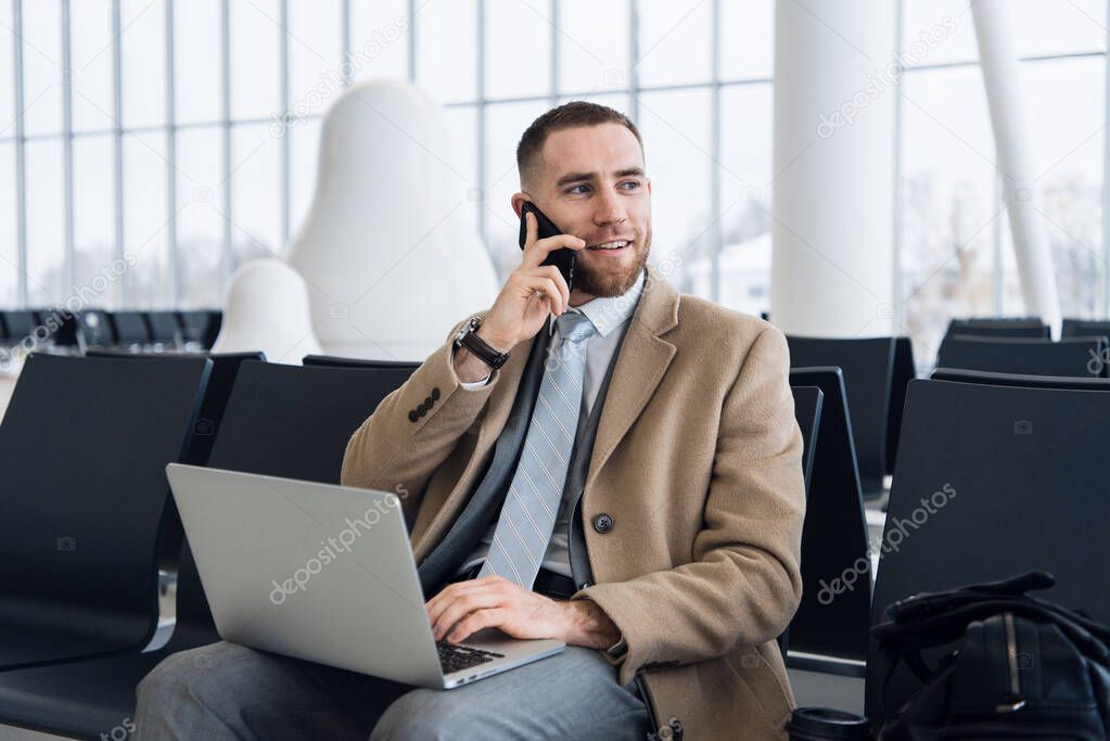 Happy businessman working on the laptop and talking on cellphone at the airport waiting lounge. Handsome caucasian businessman at waiting room in airport terminal