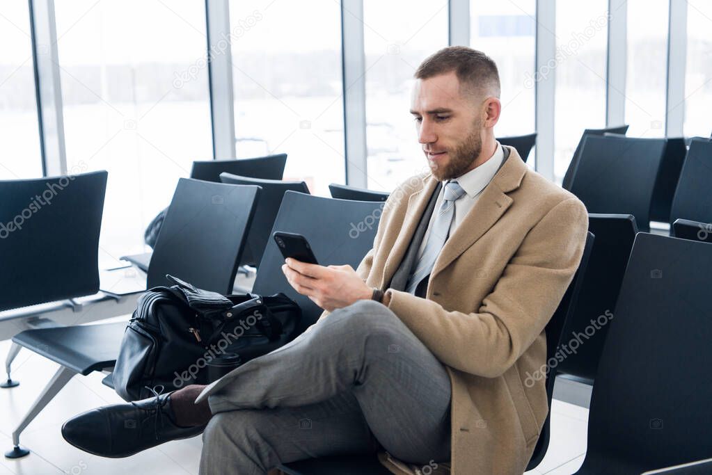 Confident man boss in luxury suit is checking e-mail via cellphone before meeting with staff. Male professional employer is typing text message on mobile phone, while is sitting in airport hallway