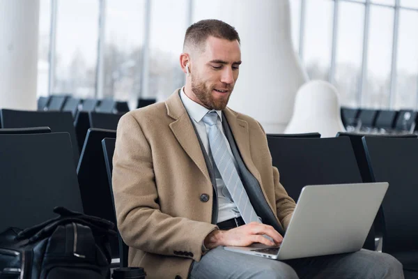 Kulaklıkla konuşan ve havaalanında dizüstü bilgisayarla çalışan yakışıklı bir iş adamı.