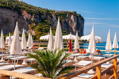 İnsanlar Napoli yakınlarındaki Vico Equensea sahil kasabasında beyaz şemsiyelerin altında dinleniyorlar. Arka planda kayalık dağlar var.