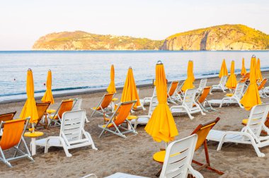 Beyaz plastik ve metal yapı, sarı kumaş güverte koltukları ve sarı şemsiyelerle birlikte Tyrhenian Denizi 'nin kumsalında, İtalya' da arka planda dağlarla birlikte.