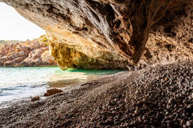 Ünlü Cala Bianca plajı manzaralı çevresindeki su üzerinde sarkan eşsiz deniz mağarasından inanılmaz deniz manzarası. Kristal berrak deniz dalgaları kayalık mağaranın içinde çakıl taşlarını yıkıyor.