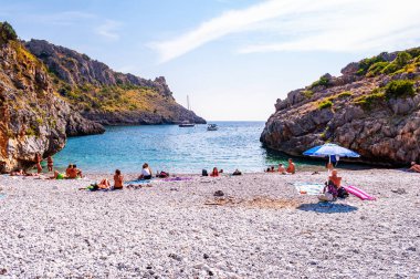 Yüzen deniz yatları, Cala Bianca sahilinde dinlenen insanlar. Suyun altında kristal berrak suyla çevrili Tyrhenian deniz koyuyla çevrili kayalar.