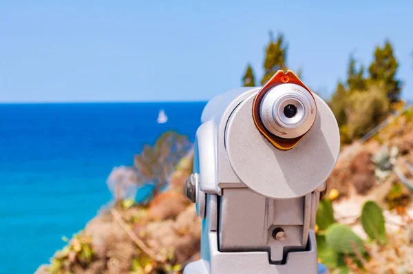 Cannocchiale turistico fisso in metallo su una gamba in piedi sul bordo di una scogliera vicino alla recinzione di tronchi di legno e osserva il mare Tirreno con yacht a vela bianco galleggiante a Tropea — Foto Stock