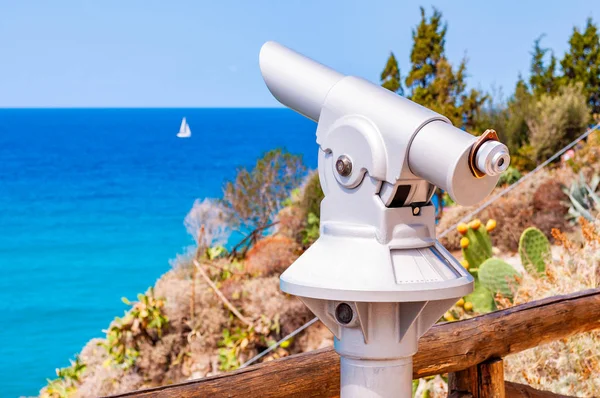 Стаціонарний туристичний телескоп, зроблений з металу на одній нозі, стоїть на краю скелі біля дерев'яного паркану колод і спостерігає за Тирренським морем з плаваючою білою вітрильною яхтою в Тропе. — стокове фото