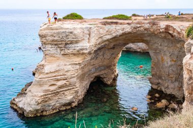 Kayanın kenarındaki turistler. Torre Sant Andrea plajı yumuşak nasırlı kayaları ve uçurumları, deniz yığınları, küçük koyları ve pürüzlü kıyı manzarasıyla
