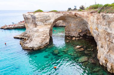 Uçurumdan atlayan, güneşlenen kayalık sahilde kristal berrak deniz suyunda yüzen kayalar, kayalıklar ve deniz yığınlarıyla Torre Sant Andrea