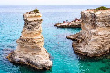 Uçurumdan atlayan, güneşlenen kayalık sahilde kristal berrak deniz suyunda yüzen kayalar, kayalıklar ve deniz yığınlarıyla Torre Sant Andrea