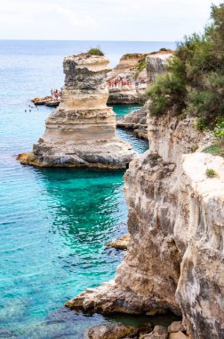 Uçurumdan atlayan, güneşlenen kayalık sahilde kristal berrak deniz suyunda yüzen kayalar, kayalıklar ve deniz yığınlarıyla Torre Sant Andrea