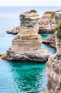 Uçurumdan atlayan, güneşlenen kayalık sahilde kristal berrak deniz suyunda yüzen kayalar, kayalıklar ve deniz yığınlarıyla Torre Sant Andrea