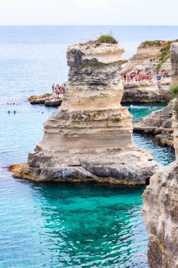 Uçurumdan atlayan, güneşlenen kayalık sahilde kristal berrak deniz suyunda yüzen kayalar, kayalıklar ve deniz yığınlarıyla Torre Sant Andrea
