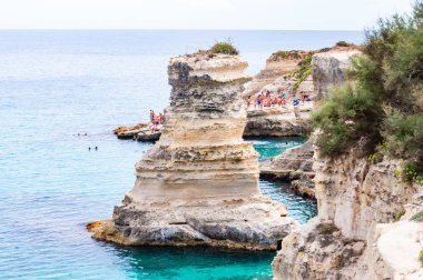 Uçurumdan atlayan, güneşlenen kayalık sahilde kristal berrak deniz suyunda yüzen kayalar, kayalıklar ve deniz yığınlarıyla Torre Sant Andrea