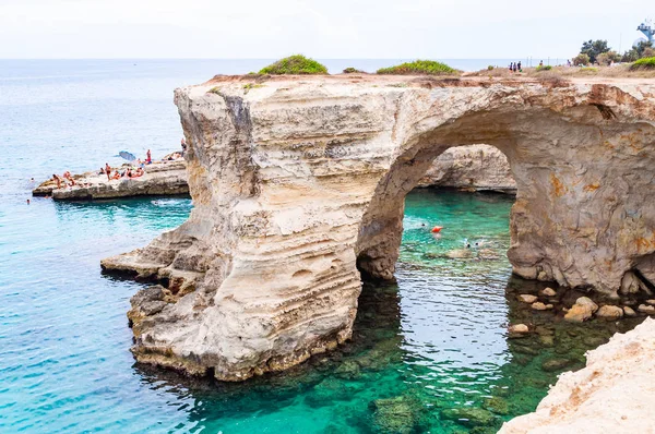 Uçurumdan atlayan, güneşlenen kayalık sahilde kristal berrak deniz suyunda yüzen kayalar, kayalıklar ve deniz yığınlarıyla Torre Sant Andrea