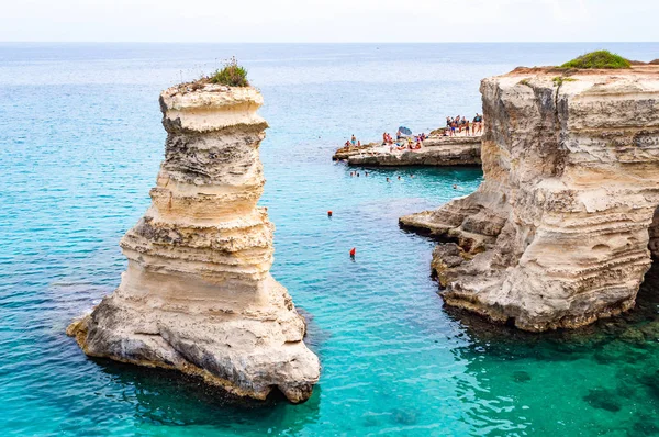 Gente buceando desde el acantilado plano, tomando el sol, nadando en aguas cristalinas en la playa rocosa Torre Sant Andrea con rocas, acantilados y pilas de mar — Foto de Stock