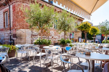 Ortaçağ Numana caddesinde çiçek açan büyük saksılarda yetişen genç zeytin ağaçları. Restoran masaları ve sandalyeler dışarıda duruyor.