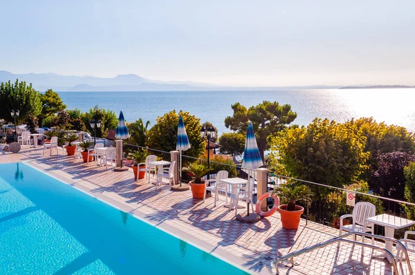 Piscina exterior con agua cristalina vibrante, sombrillas y tumbonas situadas en la costa de Garda con lago, colinas y sol de fondo Imágenes De Stock Sin Royalties Gratis