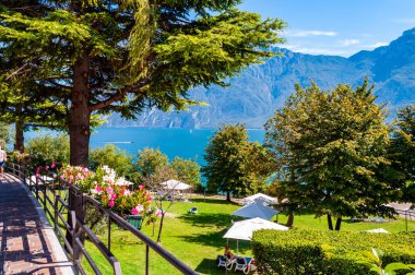 Çiçek açan Garda Gölü kıyısındaki yürüyüş yolu, yüksek yeşil ağaçlar, açık yaz çadırları ve dolomitlerle dolu yeşil çimenler.