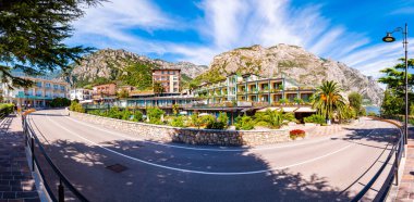 Limone Sul Garda 'nın sıcacık şehir caddesinin panoramasında büyüyen ağaçlar, arka planda muhteşem dolomite dağları olan rahat oteller.