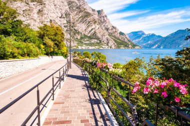Limone Sul Garda 'nın sıcacık caddesi kaldırımlı, metal korkuluklarda çiçek açan, arka planda muhteşem Garda gölü ve dolomite dağları olan ağaçlar yetiştiren. Manzara gezintisi