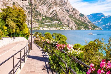 Limone Sul Garda 'nın sıcacık caddesi kaldırımlı, metal korkuluklarda çiçek açan, arka planda muhteşem Garda gölü ve dolomite dağları olan ağaçlar yetiştiren. Manzara gezintisi