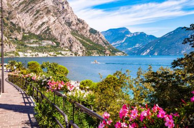 Limone Sul Garda 'nın sıcacık caddesi kaldırımlı, metal korkuluklarda çiçek açan, arka planda muhteşem Garda gölü ve dolomite dağları olan ağaçlar yetiştiren. Manzara gezintisi