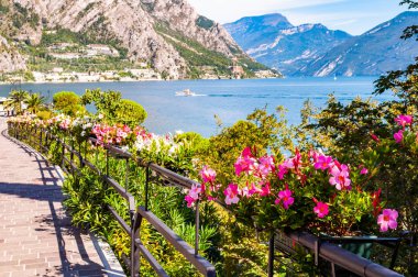 Limone Sul Garda 'nın sıcacık caddesi kaldırımlı, metal korkuluklarda çiçek açan, arka planda muhteşem Garda gölü ve dolomite dağları olan ağaçlar yetiştiren. Manzara gezintisi