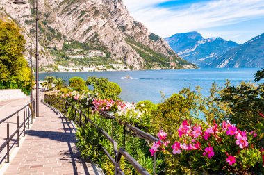 Limone Sul Garda 'nın sıcacık caddesi kaldırımlı, metal korkuluklarda çiçek açan, arka planda muhteşem Garda gölü ve dolomite dağları olan ağaçlar yetiştiren. Manzara gezintisi