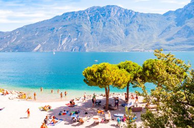 Gölün batı kıyısındaki sıcak sahil. İnsanlar yüksek yeşil çamların altında güneşlenerek yüzüyorlar. Garda, arka planda dolomitler var.
