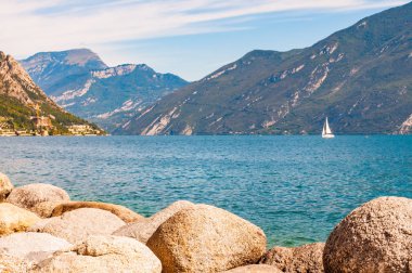 Lombardy, Kuzey İtalya 'daki güzel Garda gölünün kıyısında bulunan yuvarlak kayalar yüksek dolomit dağlarıyla çevrilidir. Gölde yüzen klasik beyaz yelkenli yat.