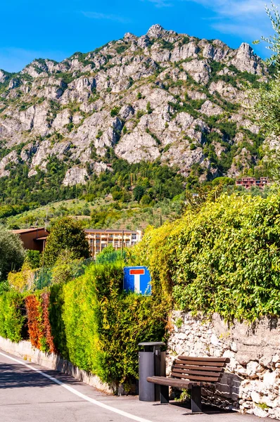 Di sisi jalan dengan bangku kayu, tanda jalan buntu, berbentuk pagar pada pagar batu dan rumah pribadi, hotel dari kota yang indah Limone Sul Garda dengan pegunungan dolomit tinggi di latar belakang — Stok Foto