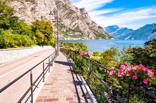 Limone Sul Garda 'nın sıcacık caddesi kaldırımlı, metal korkuluklarda çiçek açan, arka planda muhteşem Garda gölü ve dolomite dağları olan ağaçlar yetiştiren. Manzara gezintisi