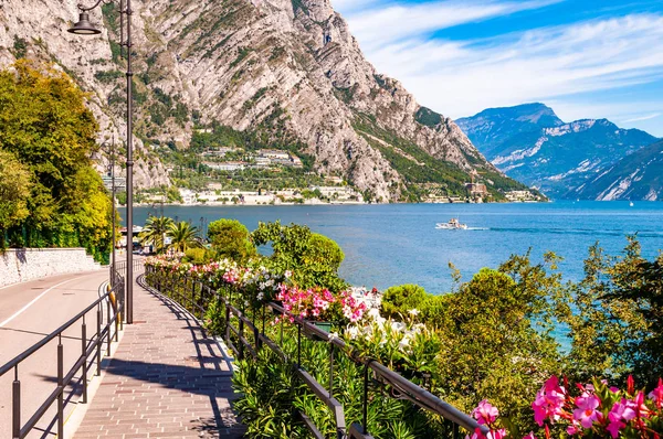 Limone Sul Garda 'nın sıcacık caddesi kaldırımlı, metal korkuluklarda çiçek açan, arka planda muhteşem Garda gölü ve dolomite dağları olan ağaçlar yetiştiren. Manzara gezintisi