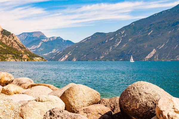 Runda stenblock ligger på stranden av vackra Gardasjön i Lombardiet, norra Italien omgiven av höga dolomitberg. Enkel klassisk vit segelbåt flyter på sjön — Stockfoto