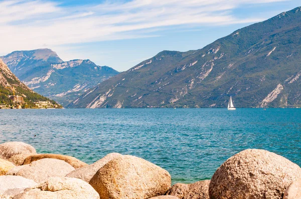 Runda stenblock ligger på stranden av vackra Gardasjön i Lombardiet, norra Italien omgiven av höga dolomitberg. Enkel klassisk vit segelbåt flyter på sjön — Stockfoto