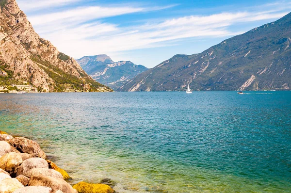 Runda stenblock ligger på stranden av vackra Gardasjön i Lombardiet, norra Italien omgiven av höga dolomitberg. Klassisk vit segelbåt flyter på sjön — Stockfoto