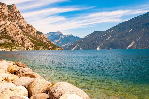 Runda stenblock ligger på stranden av vackra Gardasjön i Lombardiet, norra Italien omgiven av höga dolomitberg. Klassisk vit segelbåt flyter på sjön — Stockfoto