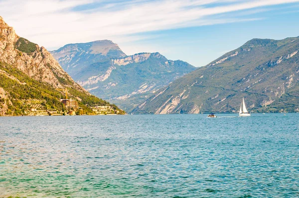 Vacker Gardasjö i Italien omgiven av höga dolomitberg. Klassisk vit segelbåt flyter på sjön. Olika hotell och privata hus byggda på den steniga stranden av sjön — Stockfoto