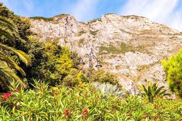 Klasická zakřivená pouliční lucerna trčící z pulzujících zelených rostlin a stromů s vysokými dolomitovými horami na pozadí v Limone Sul Garda město na západním pobřeží jezera Garda v Itálii — Stock fotografie