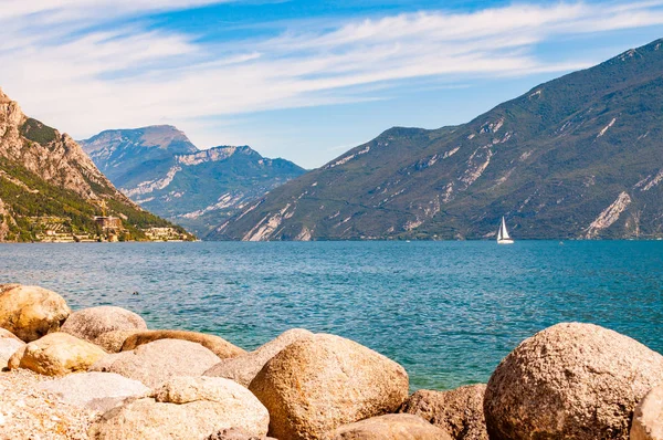 Runda stenblock ligger på stranden av vackra Gardasjön i Lombardiet, norra Italien omgiven av höga dolomitberg. Enkel klassisk vit segelbåt flyter på sjön Royaltyfria Stockbilder