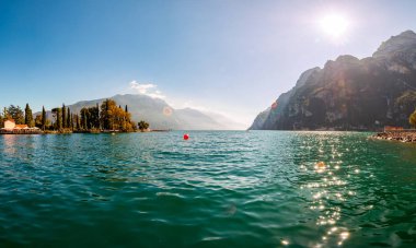 Riva del Garda şehrindeki güzel Garda Gölü kıyısındaki kayaların üzerindeki parlak güneş ışınlarına karşı panoramik manzara yüksek dolomit dağlarıyla çevrili.