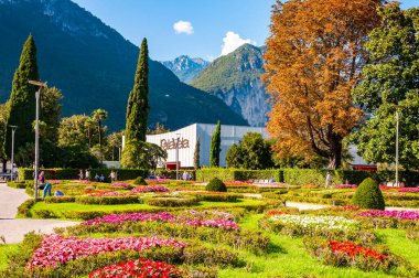 Arka planda büyüyen ve çiçek açan Palavela kongre merkeziyle Garda Gölü boyunca yürüyen insanlar.