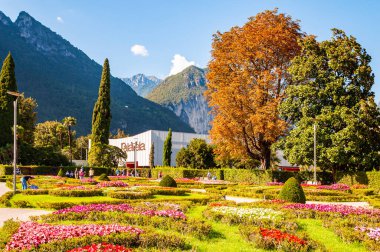 Arka planda büyüyen ve çiçek açan Palavela kongre merkeziyle Garda Gölü boyunca yürüyen insanlar.