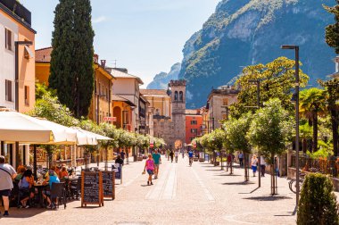 Riva del Garda 'nın manzaralı şehri. Turistler, bitkiler ve İtalyan mimarisi ile dolu rahat bir cadde. Arka planda dolomit dağları var.