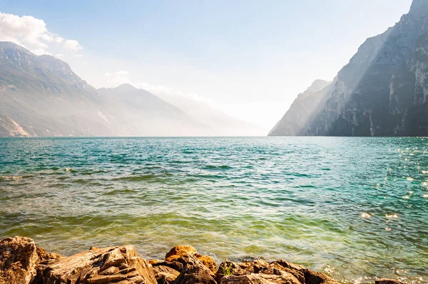 Klippiga stenar ligger på stranden av vackra Gardasjön i Lombardiet, Italien omgiven av höga dolomitberg. Solstrålar som tränger in från ovan klipporna och värmer dimmig dimma ovanför vattnet — Stockfoto